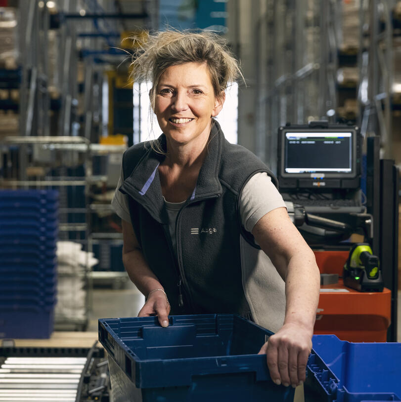 Woman in the warehouse holds a crate on the conveyor belt and smiles